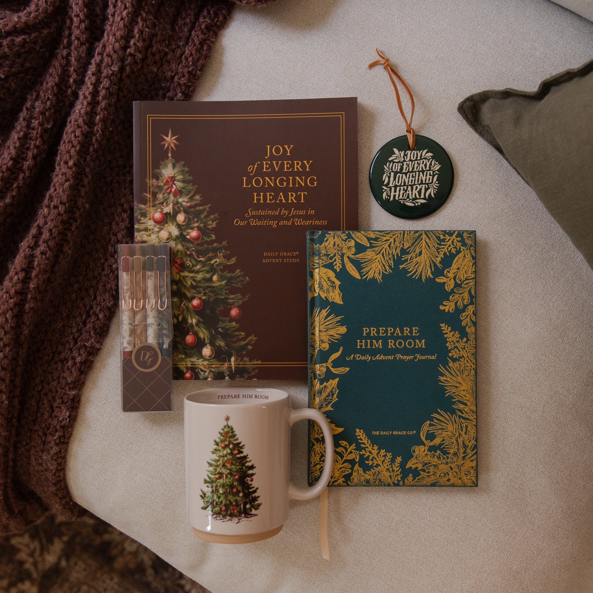 A styled holiday gift set arranged on a neutral fabric surface with a chunky knit blanket. The set includes a red book titled 'Joy of Every Longing Heart,' a teal notebook with gold botanical border and the title 'Prepare Him Room,' a white ceramic mug with a Christmas tree illustration, a small boxed set of pencils, and a round hanging ornament.