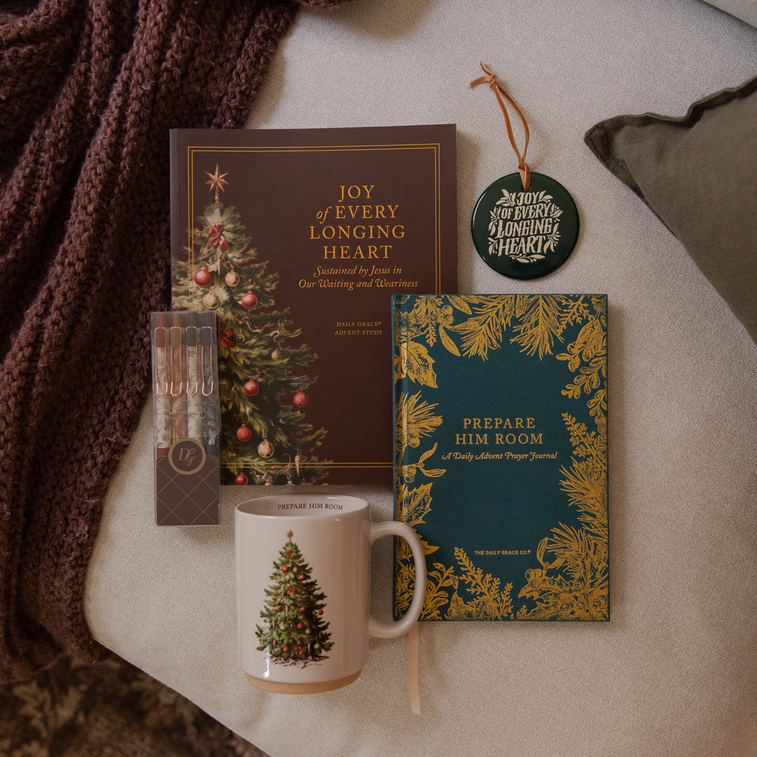 A styled holiday gift set arranged on a neutral fabric surface with a chunky knit blanket. The set includes a brown book titled 'Joy of Every Longing Heart,' a teal notebook with gold botanical border and the title 'Prepare Him Room,' a white ceramic mug with a Christmas tree illustration, a small boxed set of pencils, and a round hanging ornament.