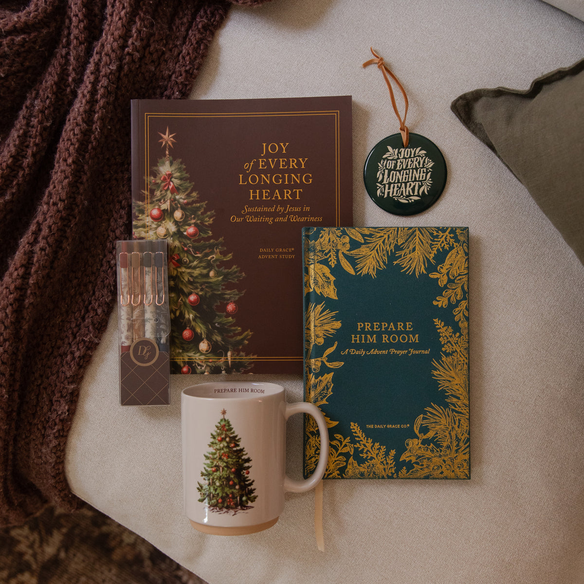 A styled holiday gift set arranged on a neutral fabric surface with a chunky knit blanket. The set includes a red book titled 'Joy of Every Longing Heart,' a teal notebook with gold botanical border and the title 'Prepare Him Room,' a white ceramic mug with a Christmas tree illustration, a small boxed set of pencils, and a round hanging ornament.