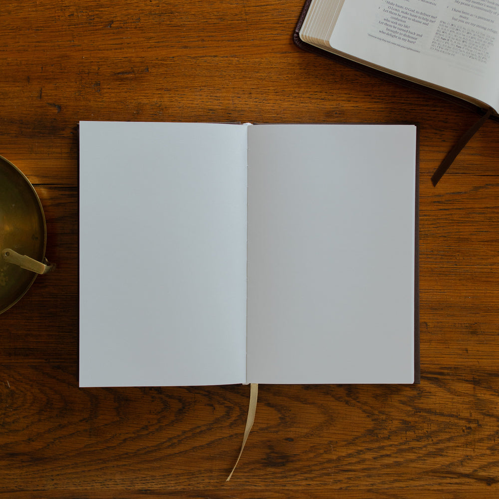 The Cross Journal opened to a blank page next to an open bible and on a wooden table