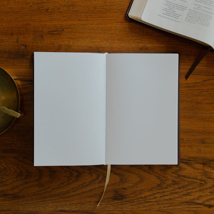 The Cross Journal opened to a blank page next to an open bible and on a wooden table