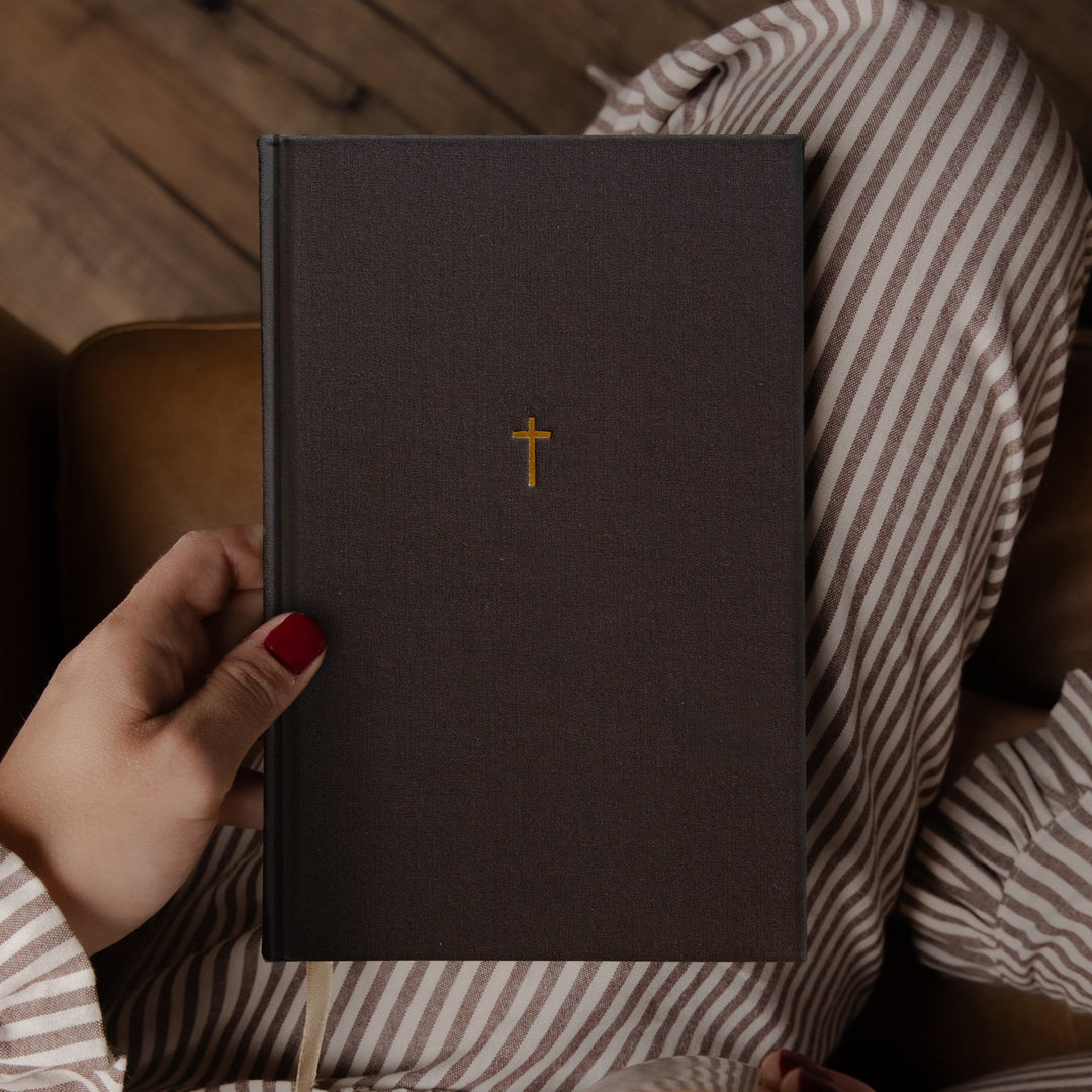 A person in striped pajamas holds a dark brown hardcover journal with a small gold cross on the cover, photographed from above while sitting on a brown leather couch with a coffee mug in the foreground.
