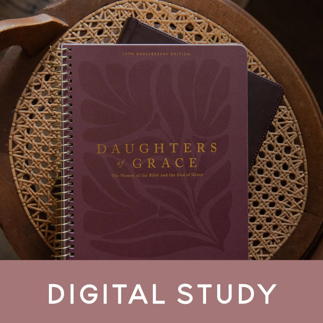 A spiral-bound book titled "Daughters of Grace" in a deep burgundy cover with gold lettering, placed on a woven round tray. A dark bookmark or card is tucked behind it.