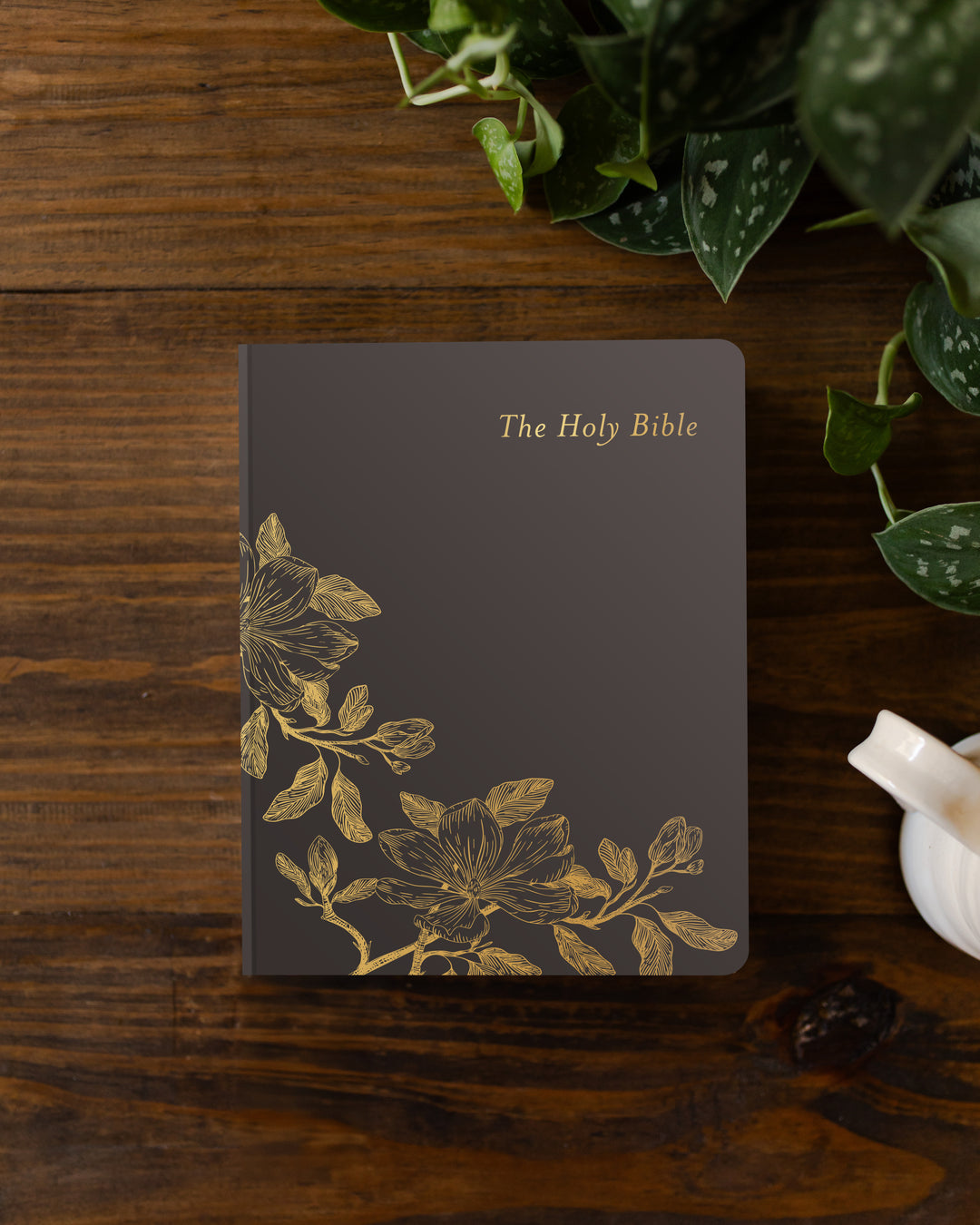 Dark brown Bible with gold floral design on a wooden surface with plants.