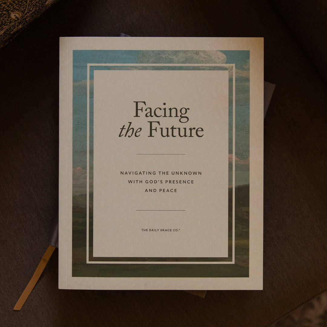 A book titled 'Facing the Future' lies on a dark brown leather surface, surrounded by patterned fabric and a small wooden stick. The cover is cream with a teal and green border design.