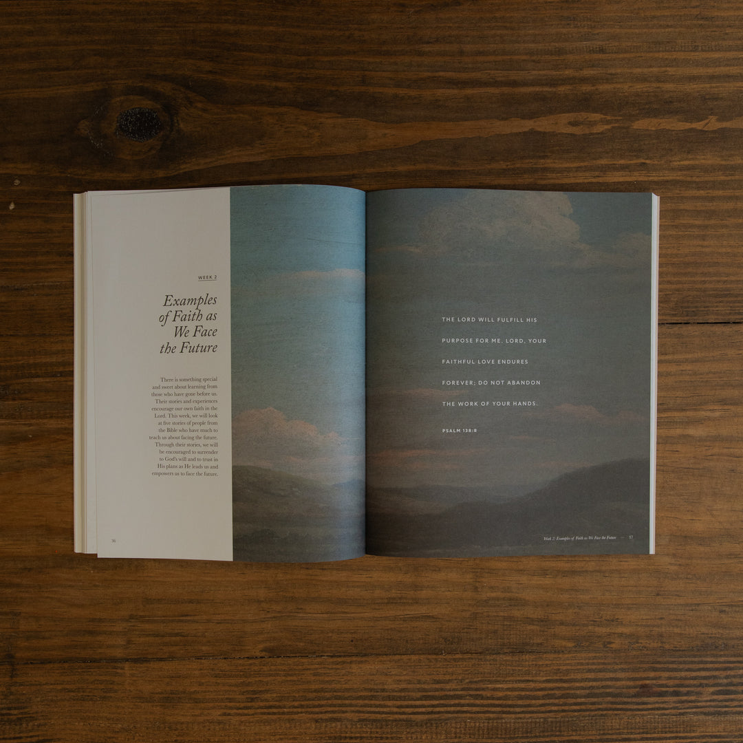 An open book laid flat on a dark wood surface, showing a two-page spread with a landscape photograph of mountains and clouds across the center and serif text on the left and right pages.