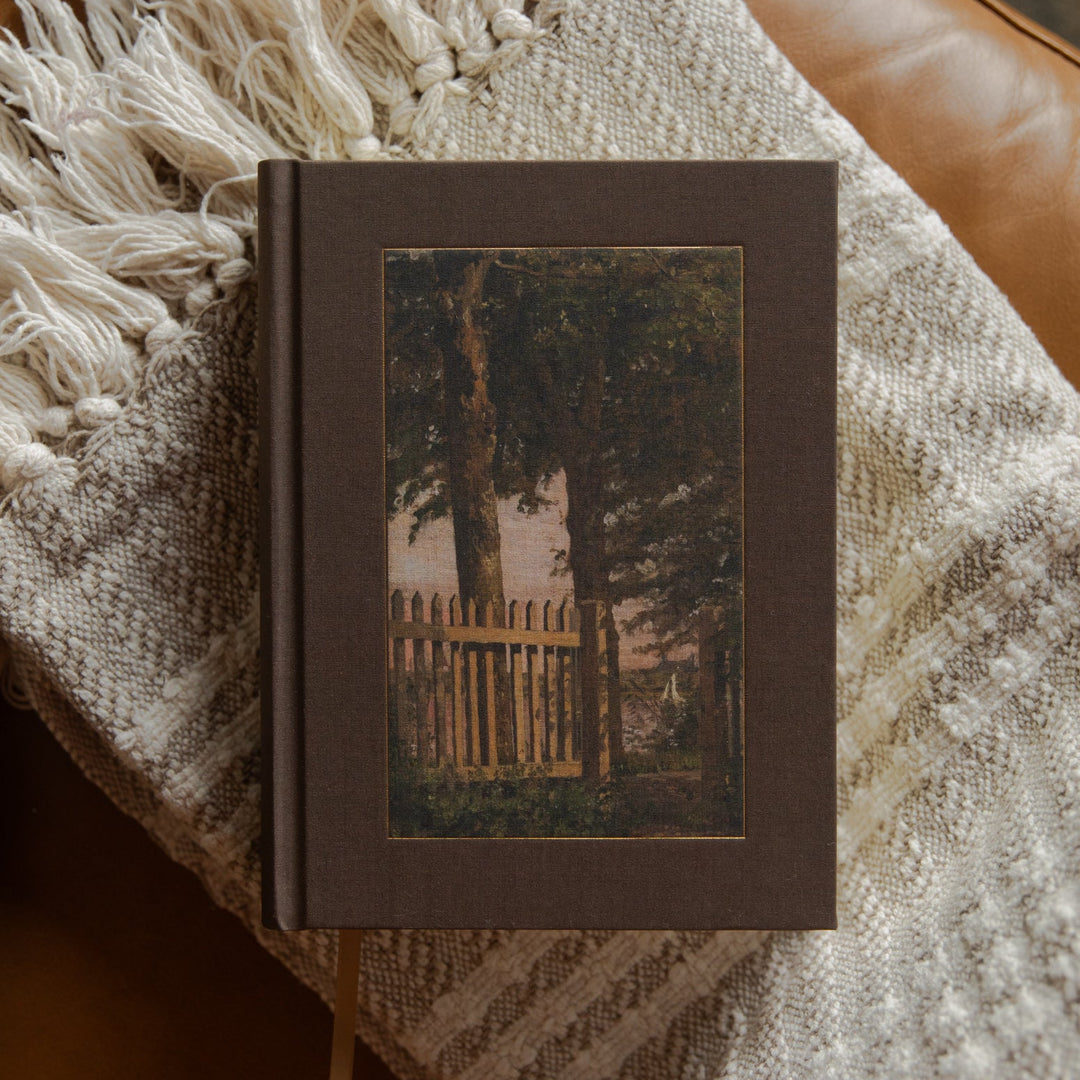 A hardcover bible with a dark brown cover and a central vintage-style illustration of a tree and picket fence, resting on a cream knit throw blanket with fringe on a brown leather couch.