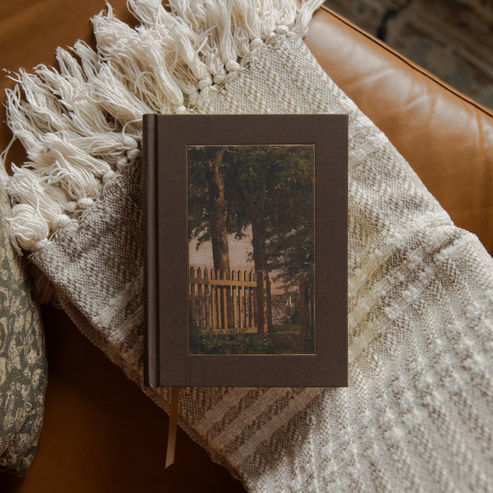 A hardcover bible with a dark brown cover and a central vintage-style illustration of a tree and picket fence, resting on a cream knit throw blanket with fringe on a brown leather couch.