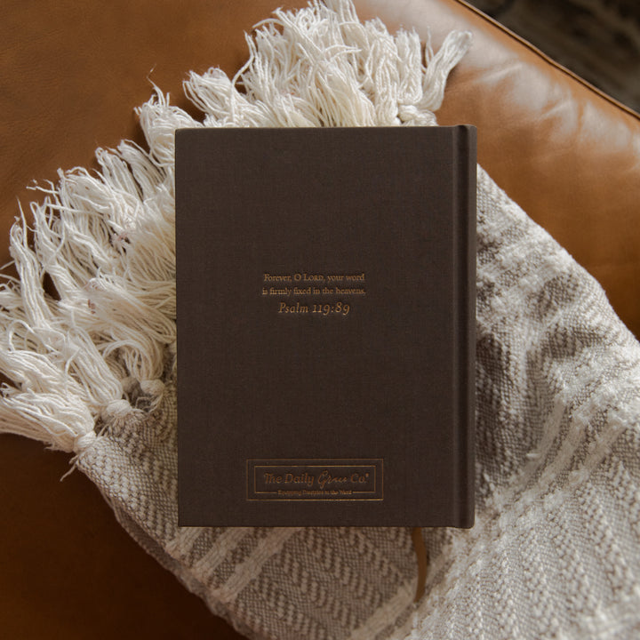 A dark brown hardcover bible from The Daily Green Co, featuring gold foil text, styled on a beige and white patterned fringed throw blanket over a brown leather couch.