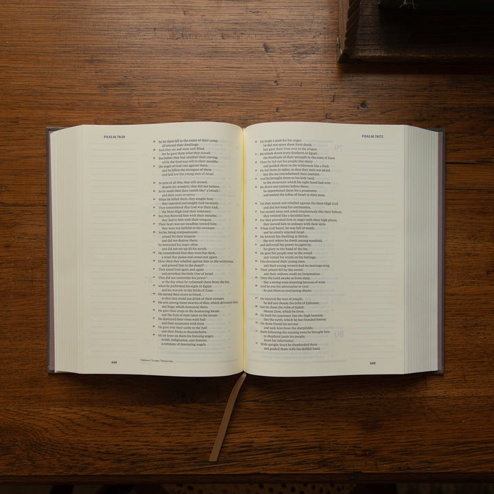 An open bible with printed text spread across two pages, placed on a dark wooden tabletop. A bookmark ribbon hangs from the bottom center.
