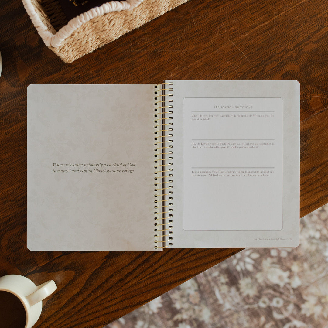 Flourishing in Motherhood spiral study opened to the application and questions page on a wooden table near a coffee cup