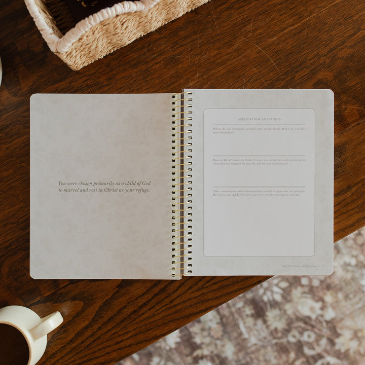 Flourishing in Motherhood spiral study opened to the application and questions page on a wooden table near a coffee cup