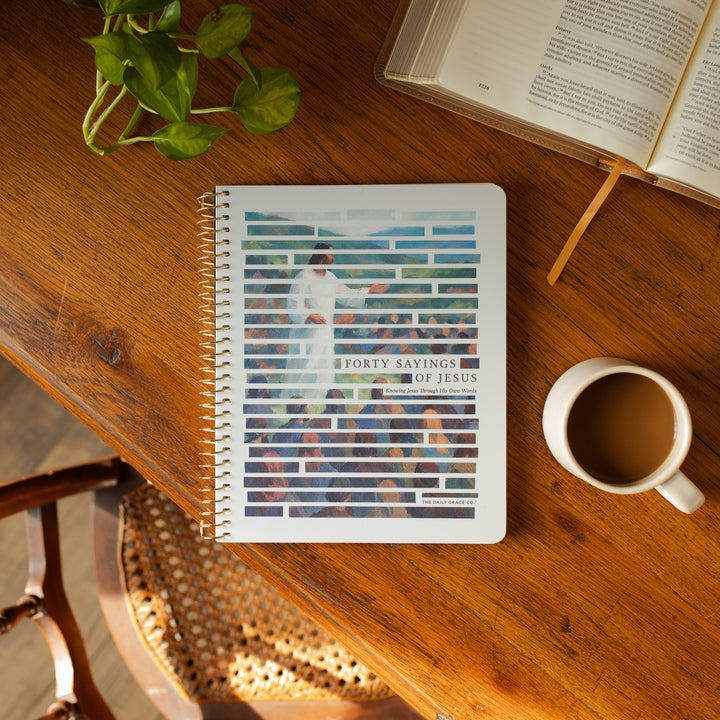 The Forty Sayings of Jesus Spiral cover on a wooden table next to a white coffee cup and an open bible