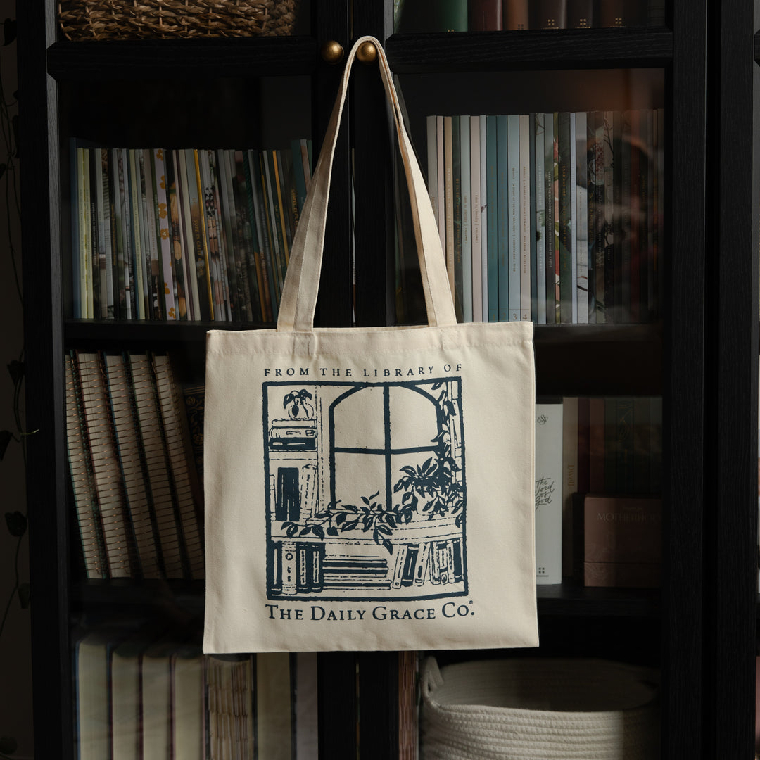 An off-white canvas tote bag with dark blue printed graphics and text hanging from a gold hook on a black bookshelf filled with books. The bag features the text 'FROM THE LIBRARY OF THE Daily Grace Co.