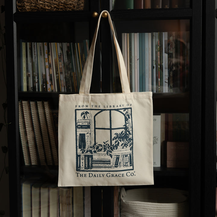 An off-white canvas tote bag with dark blue printed graphics and text hanging from a gold hook on a black bookshelf filled with books. The bag features the text 'FROM THE LIBRARY OF THE Daily Grace Co.