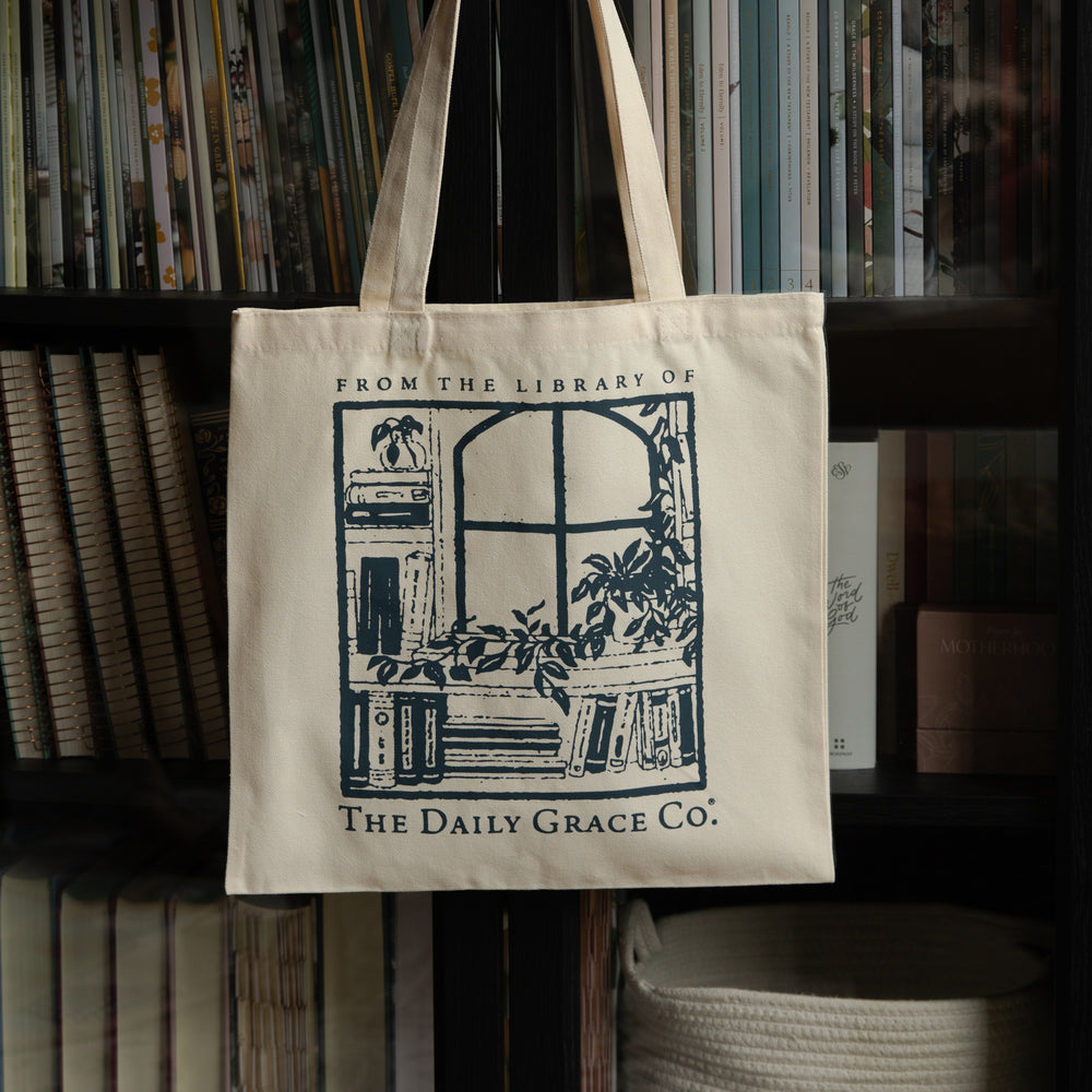 An off-white canvas tote bag with a dark blue graphic print of a library scene and the text 'FROM THE LIBRARY OF THE DAILY GRACE CO.' hanging in front of a bookshelf.