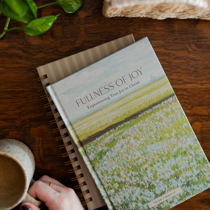 The Fullness of Joy cover on a closed journal on a wooden table next to a coffee cup with someone's hand on it