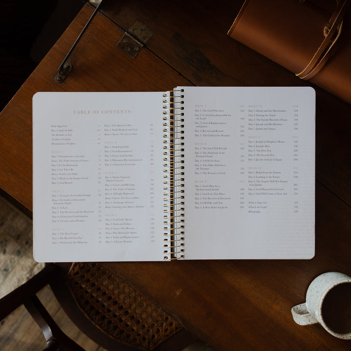 Genesis Spiral opened to the table of contents page on a wooden table near a coffee cup