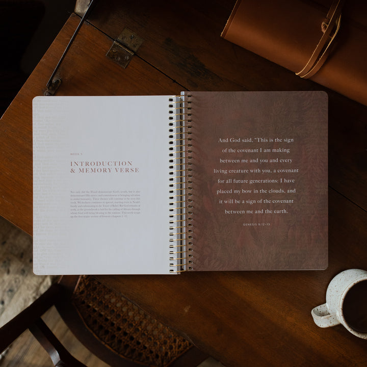 Genesis Spiral opened to the week 5 introduction page on a wooden table near a coffee cup