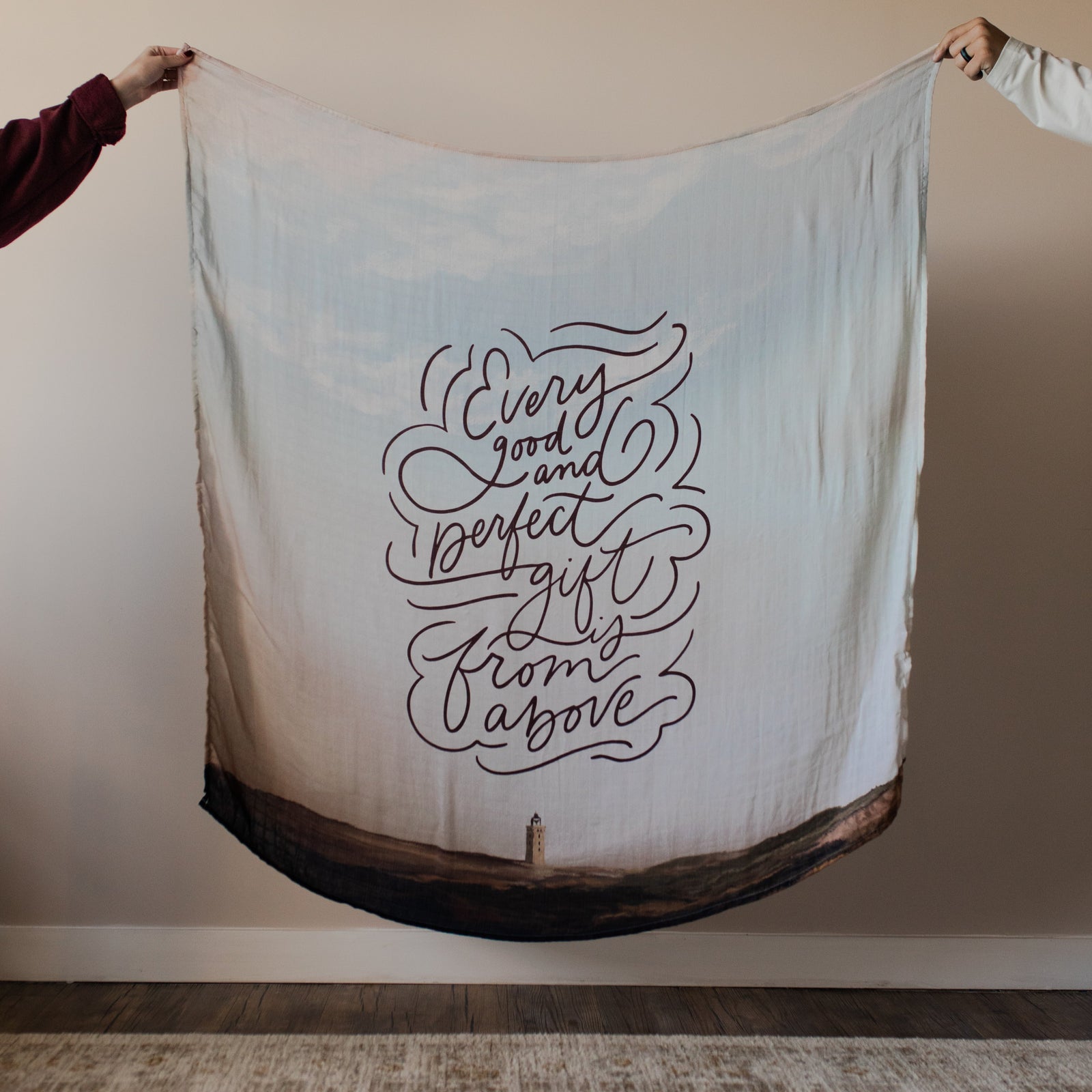 Good and Perfect Gift Swaddle held up by two hands near a wall