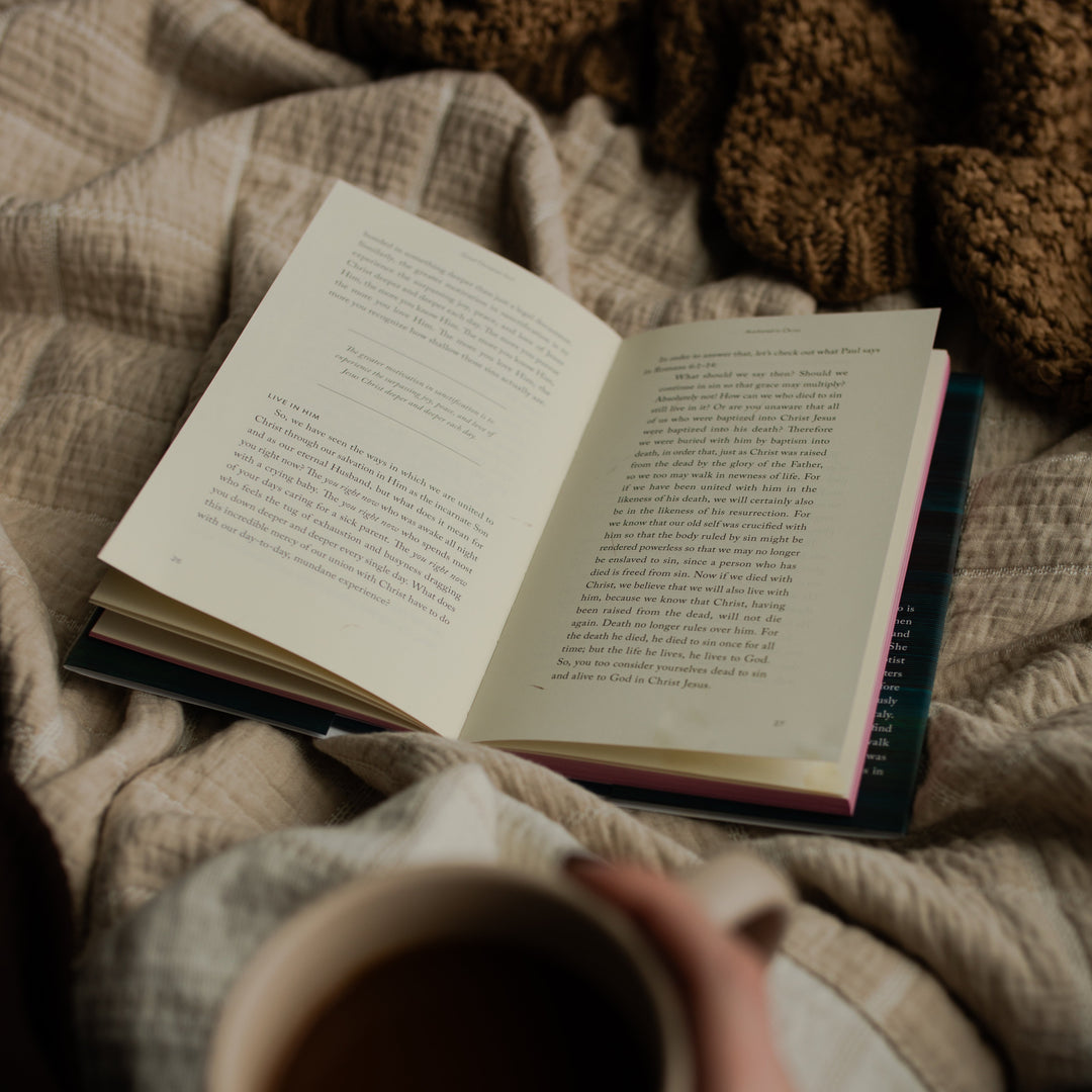 The Good Christian Girl book opened to a random page in the book on a tan blanket near a woman's hand holding a coffee cup