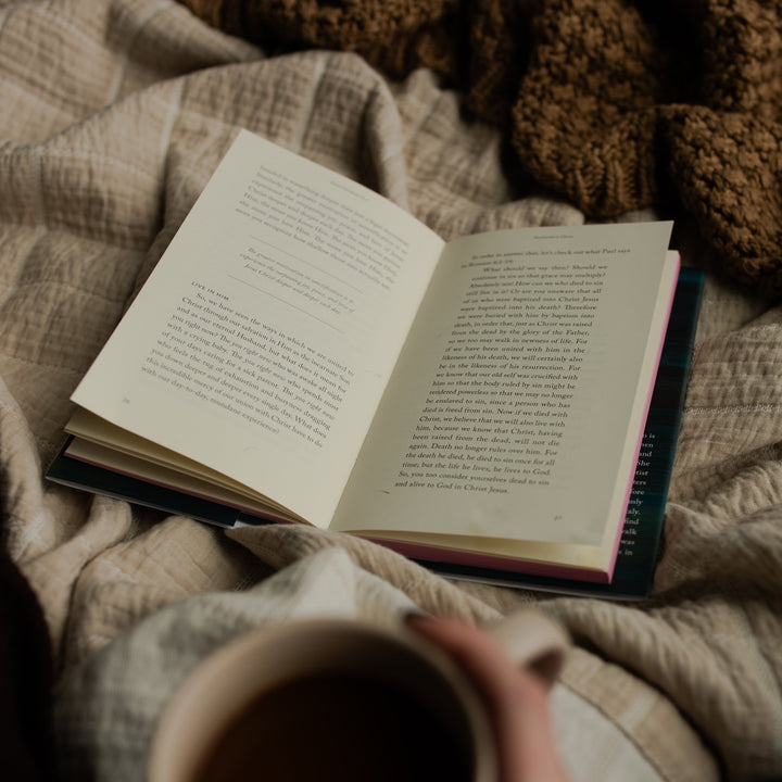 The Good Christian Girl book opened to a random page in the book on a tan blanket near a woman's hand holding a coffee cup