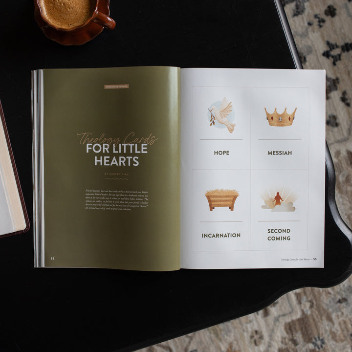 Gospel at Home Issue 10 opened to the Theology Cards for Little Hearts page on a black table near a coffee cup
