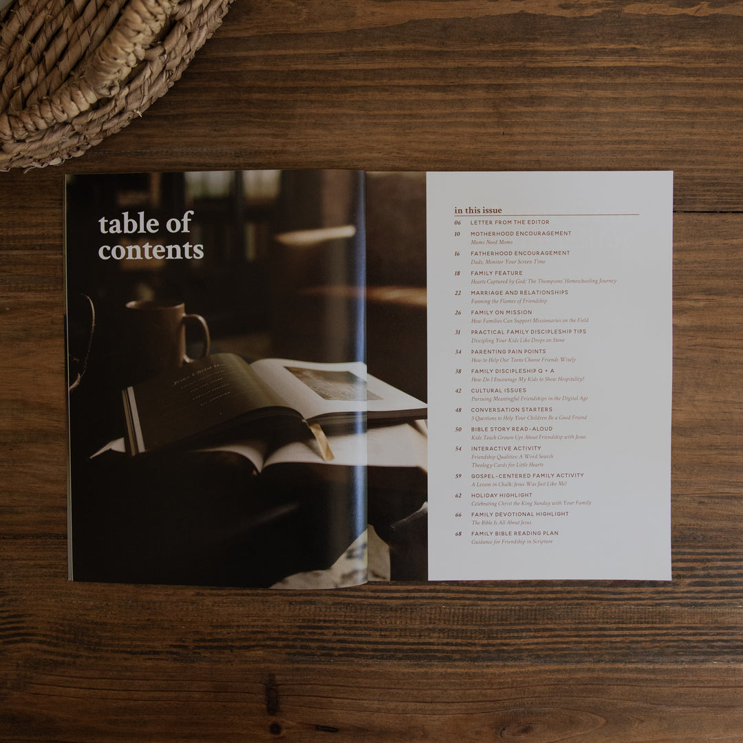 An open printed magazine spread lying on a wooden table, showing a table of contents page with a dark photo of books and a mug on the left and a list of chapters on the right.