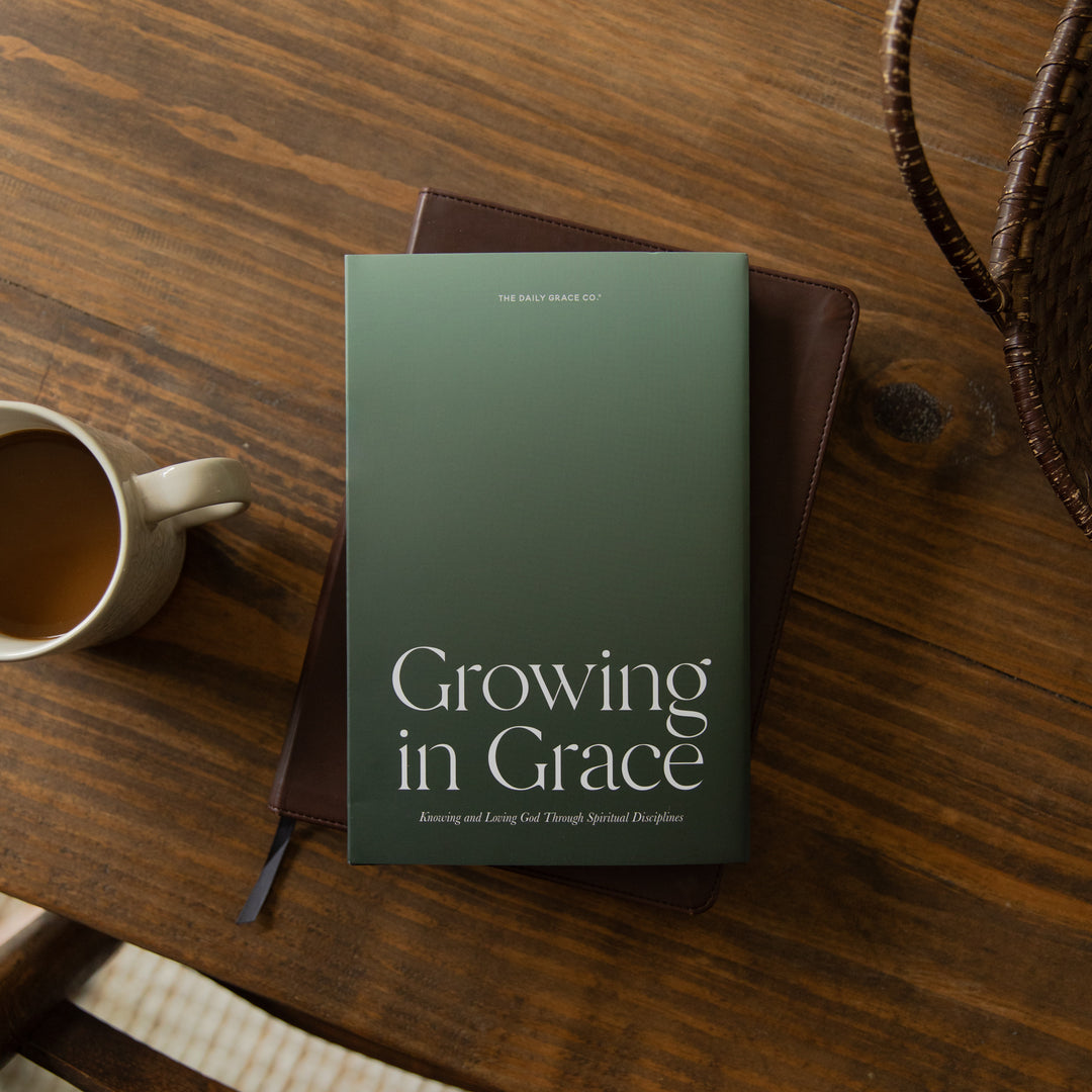 A hardcover book titled 'Growing in Grace' by Dr. Mark Deas lies on a dark brown leather journal, styled on a rustic wooden table next to a white ceramic coffee mug.