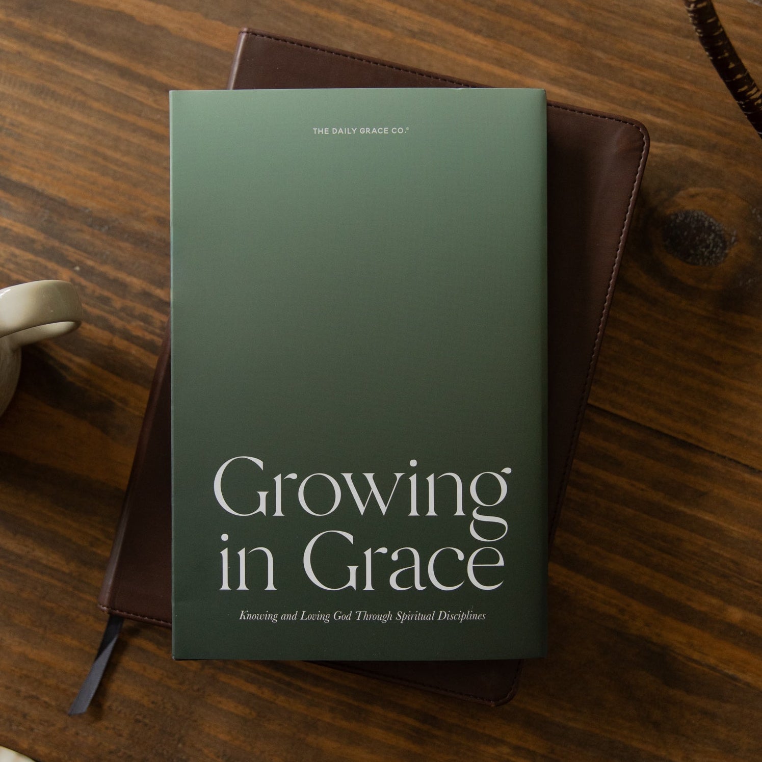 A hardcover book titled 'Growing in Grace' by Dr. Mark Deas lies on a dark brown leather journal, styled on a rustic wooden table next to a white ceramic coffee mug.