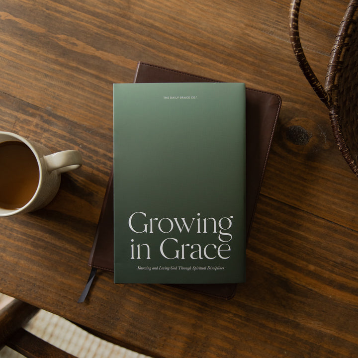 A hardcover book titled 'Growing in Grace' by Dr. Mark Deas lies on a dark brown leather journal, styled on a rustic wooden table next to a white ceramic coffee mug.