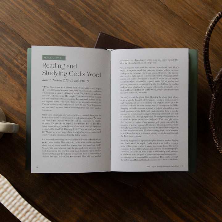 An open Bible book laid flat on a dark wooden table, showing two spread pages with printed text. A dark cloth or book lies partially underneath, and a woven basket edge is visible at the right.