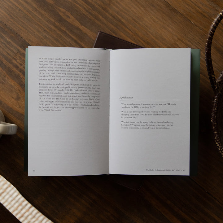 An open book laid flat on a dark wooden table, showing two printed interior pages with text and a small section titled 'Application.' A dark cover or folder is partially visible behind the open pages, and part of a woven strap and a light object appear at the edges.