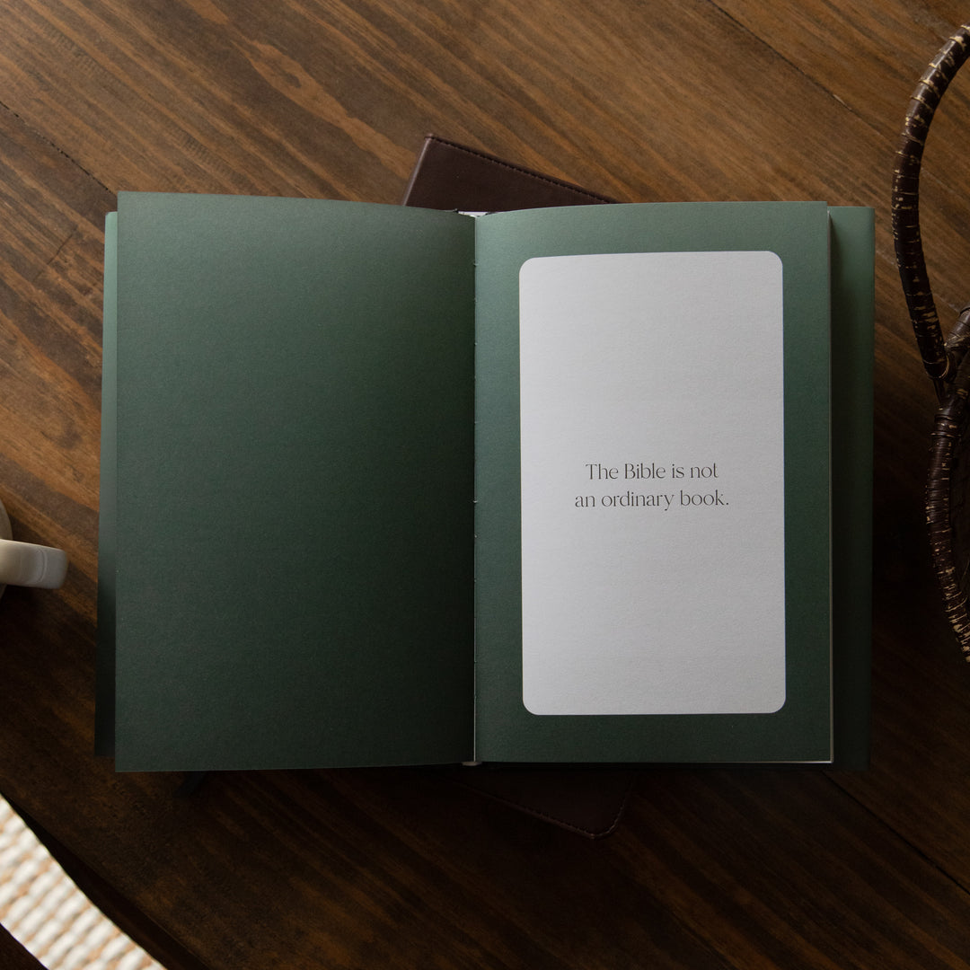 An open dark green book lying on a wooden table, with a white page inside that reads, 'The Bible is not an ordinary book.'