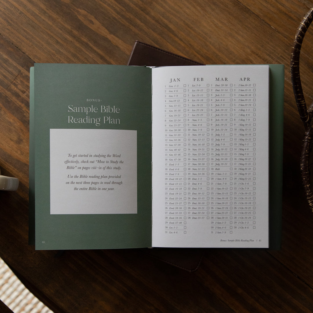 An open Bible reading plan booklet laid on a dark wooden table, showing a green cover page with the title 'Sample Bible Reading Plan' and an interior spread with a monthly calendar grid. A brown leather strap and a small woven textile are partially visible around it.