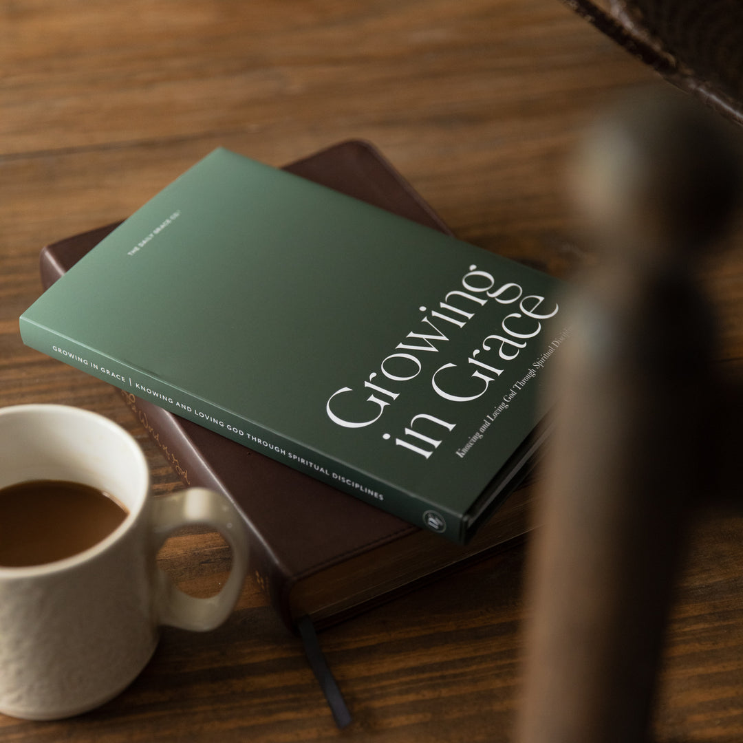 A green book titled 'Growing in Grace' resting on a brown book on a wooden table, with a white mug of coffee in the foreground and a blurred chair back on the right.
