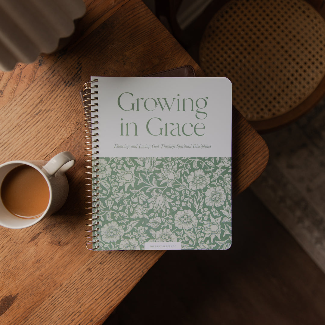 A spiral-bound notebook titled 'Growing in Grace' lies on a wooden table next to a white mug filled with coffee. The notebook cover is white on the top half with green text, and the bottom half features a light green floral pattern.