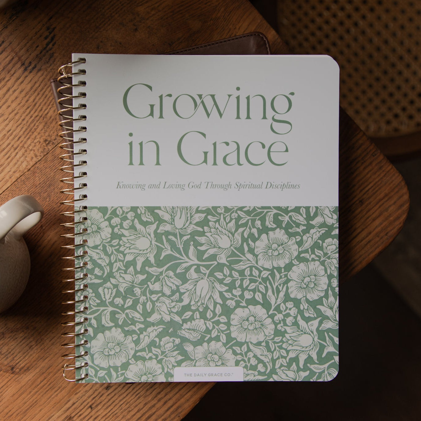 A spiral-bound notebook titled 'Growing in Grace' lies on a wooden table next to a white mug filled with coffee. The notebook cover is white on the top half with green text, and the bottom half features a light green floral pattern.