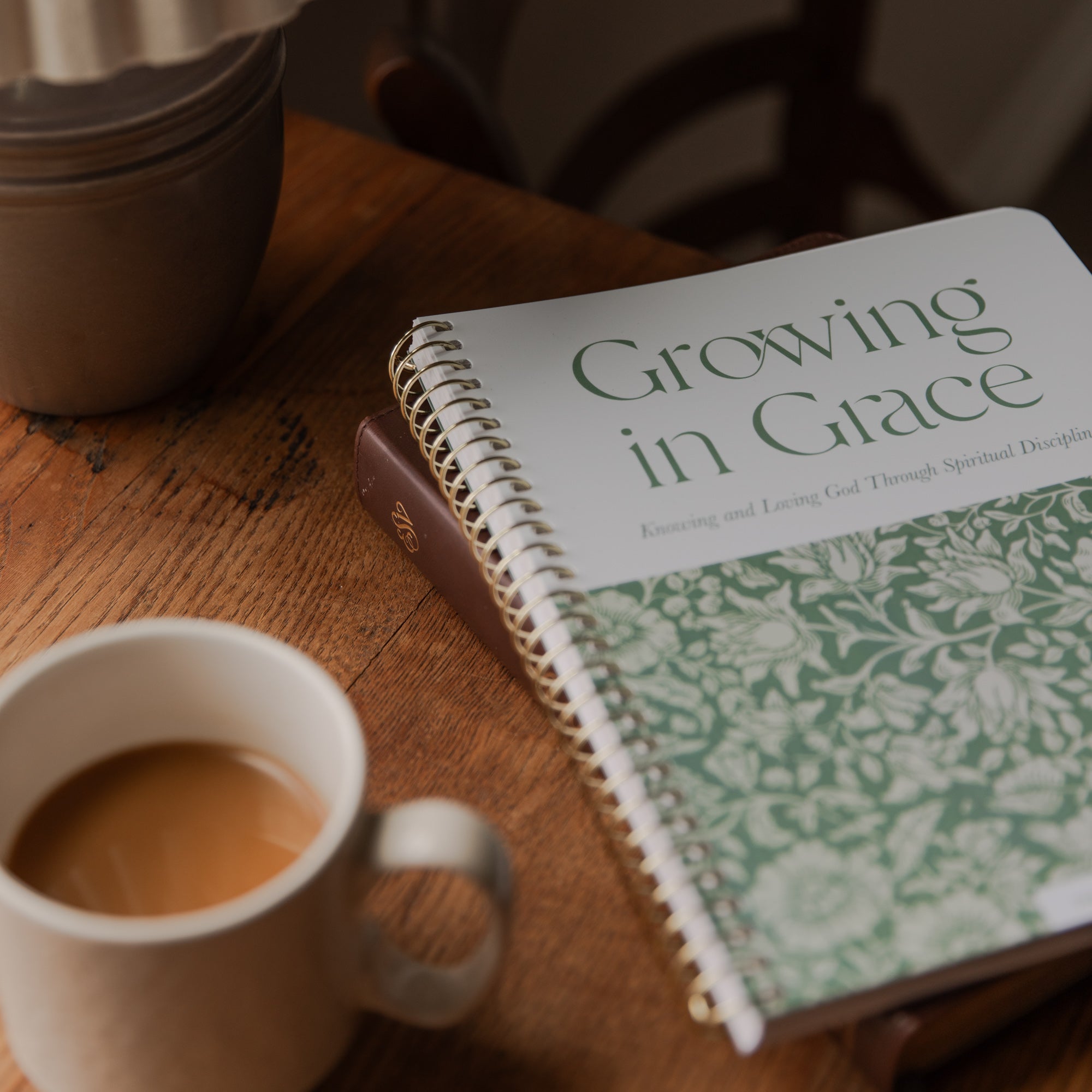 A spiral-bound book titled 'Growing in Grace' lies open on a wooden table, partially covered by a white ceramic mug filled with coffee and another mug in the background.