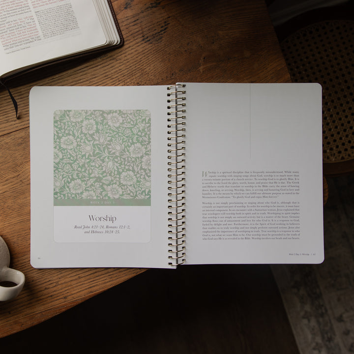 An open spiral-bound book or journal lies on a wooden table, showing a floral patterned page on the left and a text page on the right. A coffee cup is partially visible at the lower left, and another open book is at the upper left.