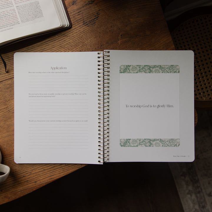 An open spiral-bound prayer journal lies on a wooden table, showing lined application text on the left page and a blank prayer page with decorative green floral bands on the right. An open book and part of a cup are visible around it.