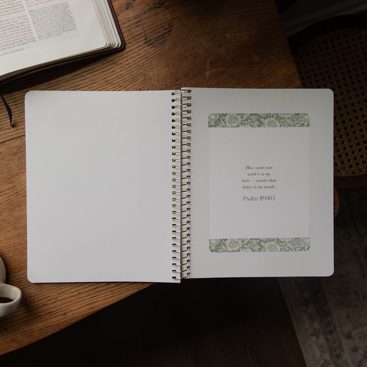An open spiral-bound journal or notebook resting on a wooden table. The left page is blank, and the right page features a centered white card with a Bible verse from Psalm 19:28 printed in black text, framed by two narrow green floral patterned borders at the top and bottom.