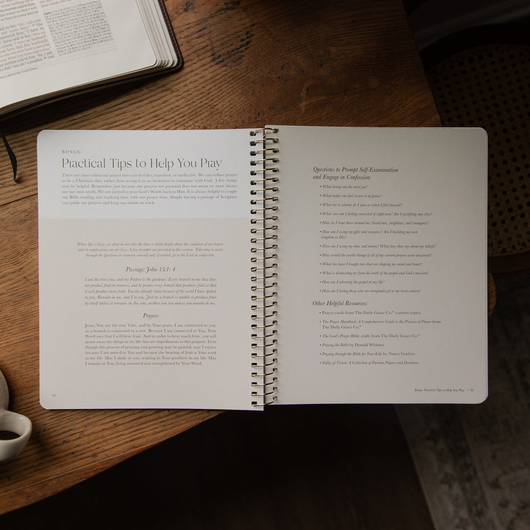 An open spiral-bound booklet or notebook on a wooden table, showing printed text pages. Another open book is partially visible at the top left, and a small cup edge appears at the bottom left.