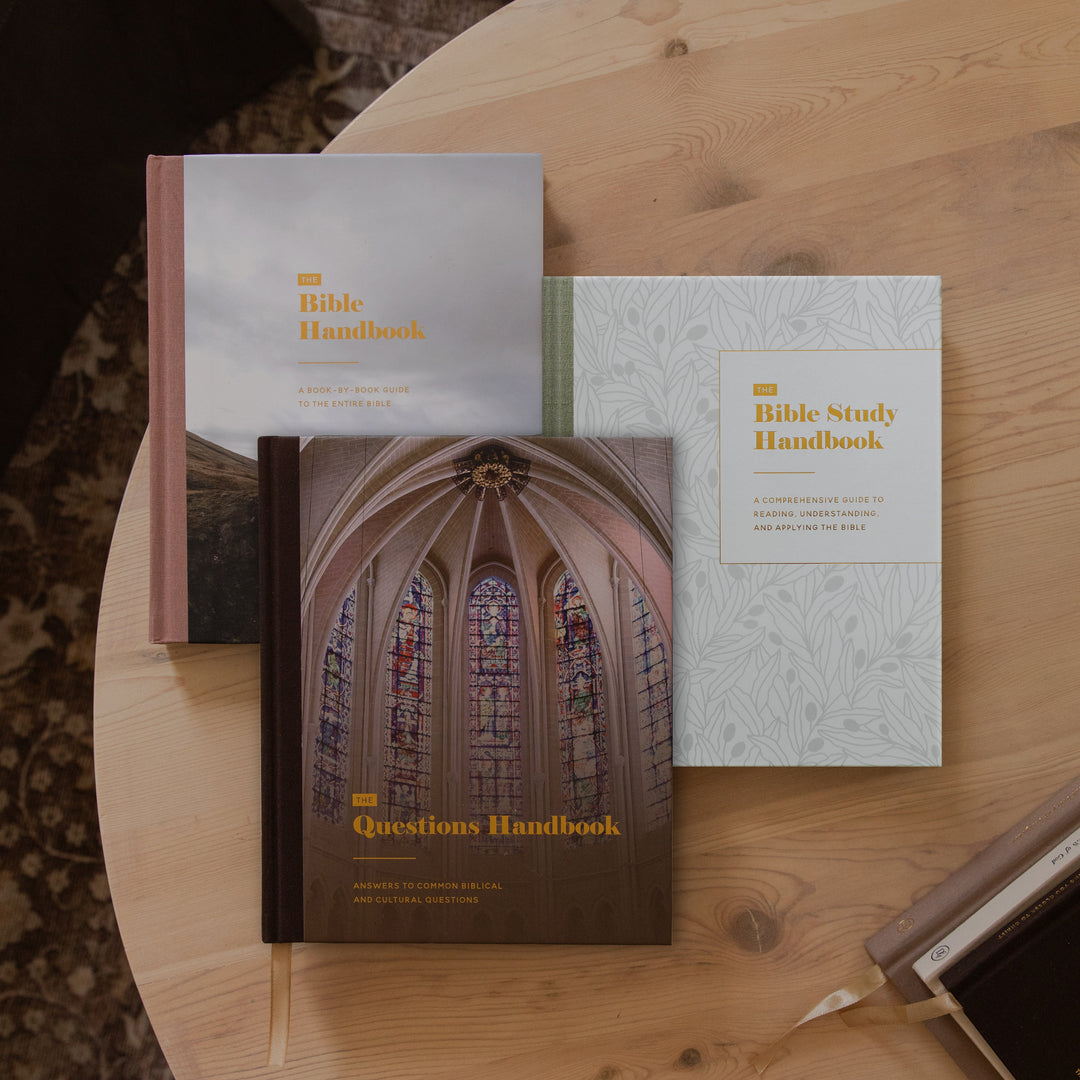 Three Bible handbooks arranged on a round wooden table: the Bible Handbook, the Questions Handbook, and the Bible Study Handbook. A pen and another book are partially visible nearby.
