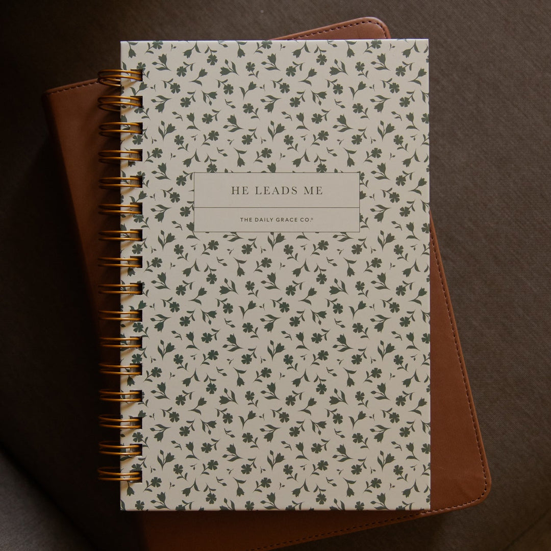 A spiral-bound journal with a white cover covered in a small green floral pattern and a tan label reading 'HE LEADS ME', resting on a brown faux-leather notebook on a dark gray upholstered chair.