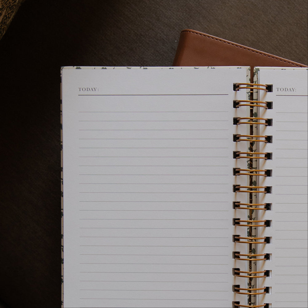 An open spiral-bound planner or notebook with lined pages, photographed on a dark brown textured surface. A brown leather-like cover edge is visible at the top, and the gold-toned spiral binding runs vertically down the center.