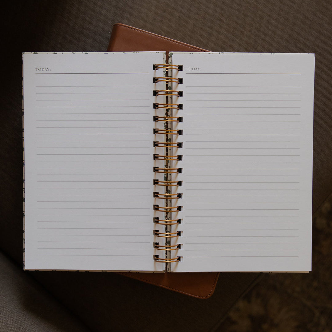 An open spiral-bound lined notebook with a gold-toned coil, photographed from above on a brown upholstered chair.