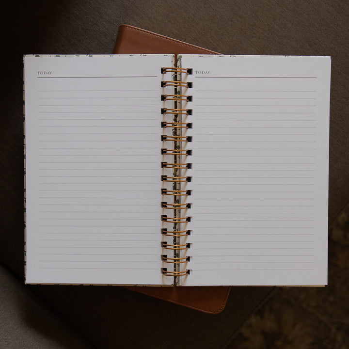 An open spiral-bound lined notebook with a gold-toned coil, photographed from above on a brown upholstered chair.