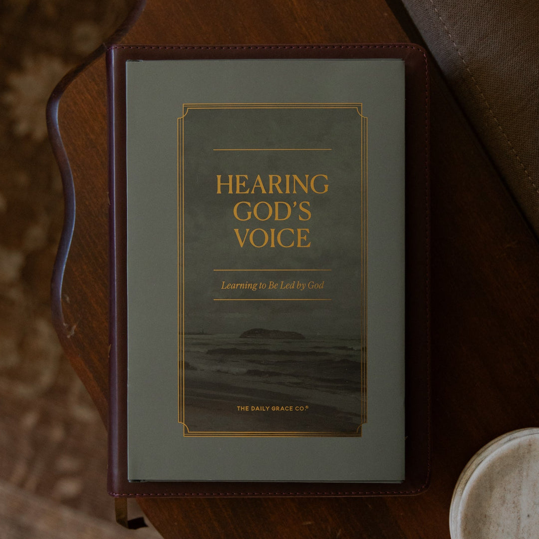 A book titled 'HEARING GOD'S VOICE' resting on a dark wooden table next to a small white dish. The book cover is a muted sage green with gold lettering and a thin gold border.