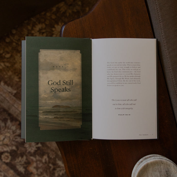 An open book titled 'God Still Speaks' resting on a dark wooden table, with a plate partially visible in the foreground and a patterned rug in the background.